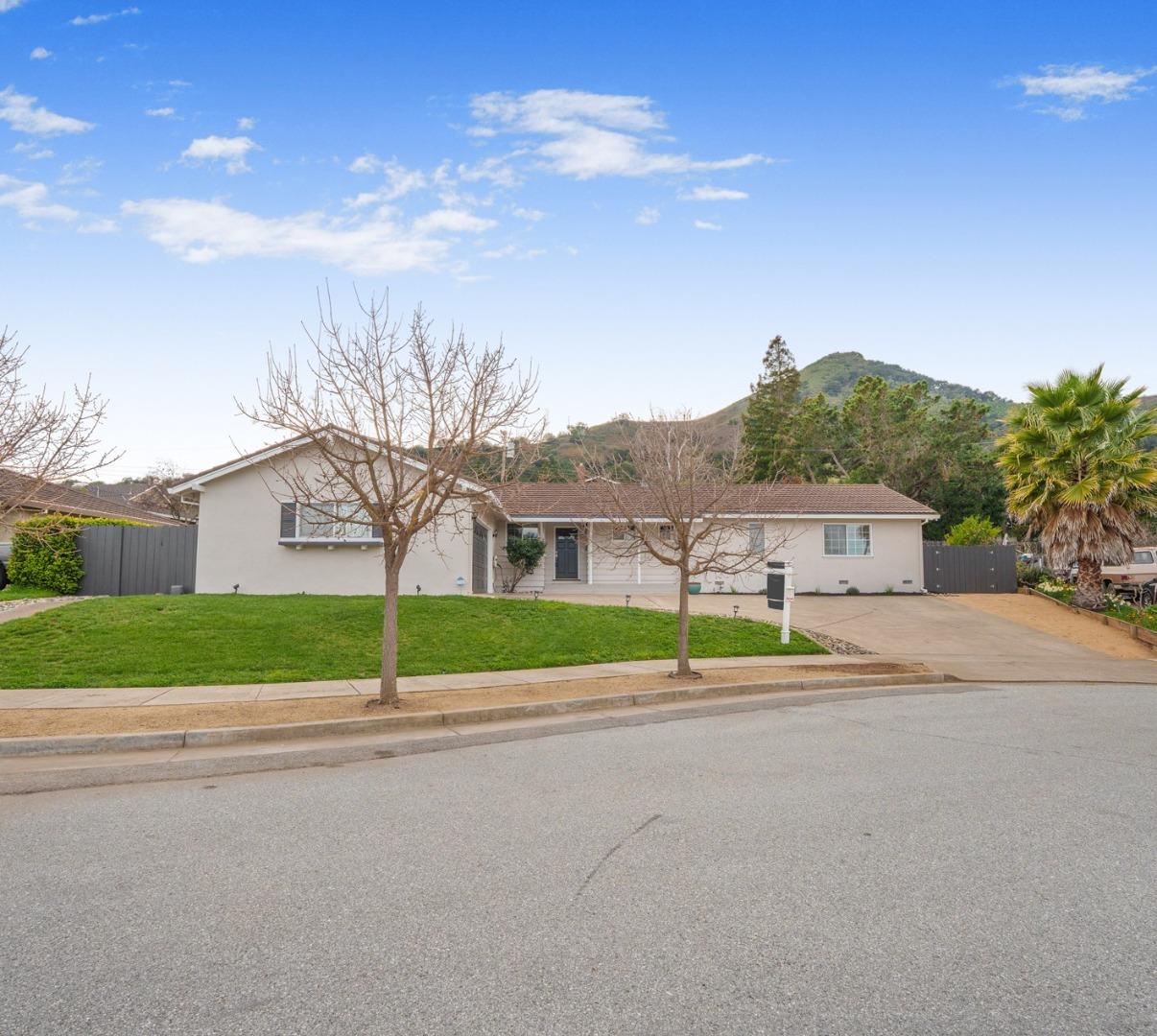 Single-story home with a front lawn, wide driveway, and green hillside behind it.