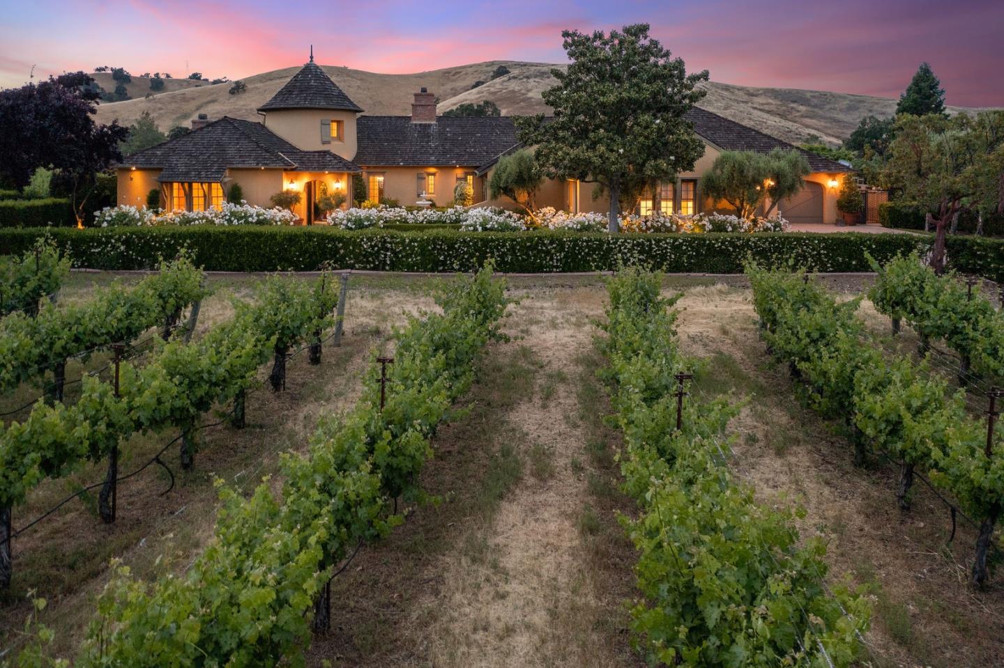 Large tiled-roof home behind a hedge, with vineyard rows and rolling hills at dusk.