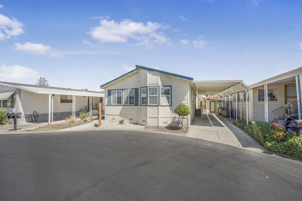 Light-colored manufactured home with a covered carport beside a paved community street.