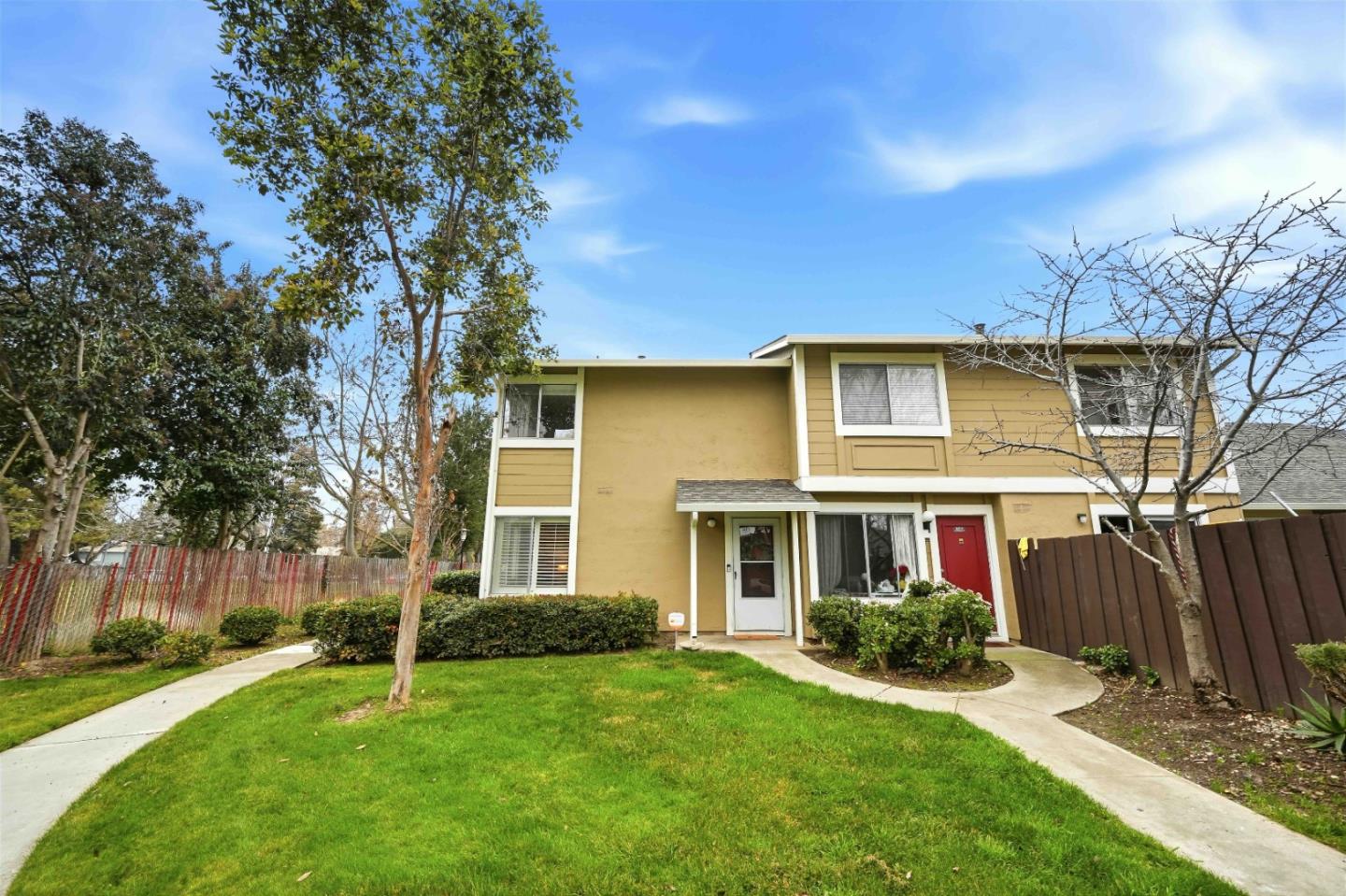 Two-story tan townhouse with white trim and a red door, beyond a lawn and curved walkways.