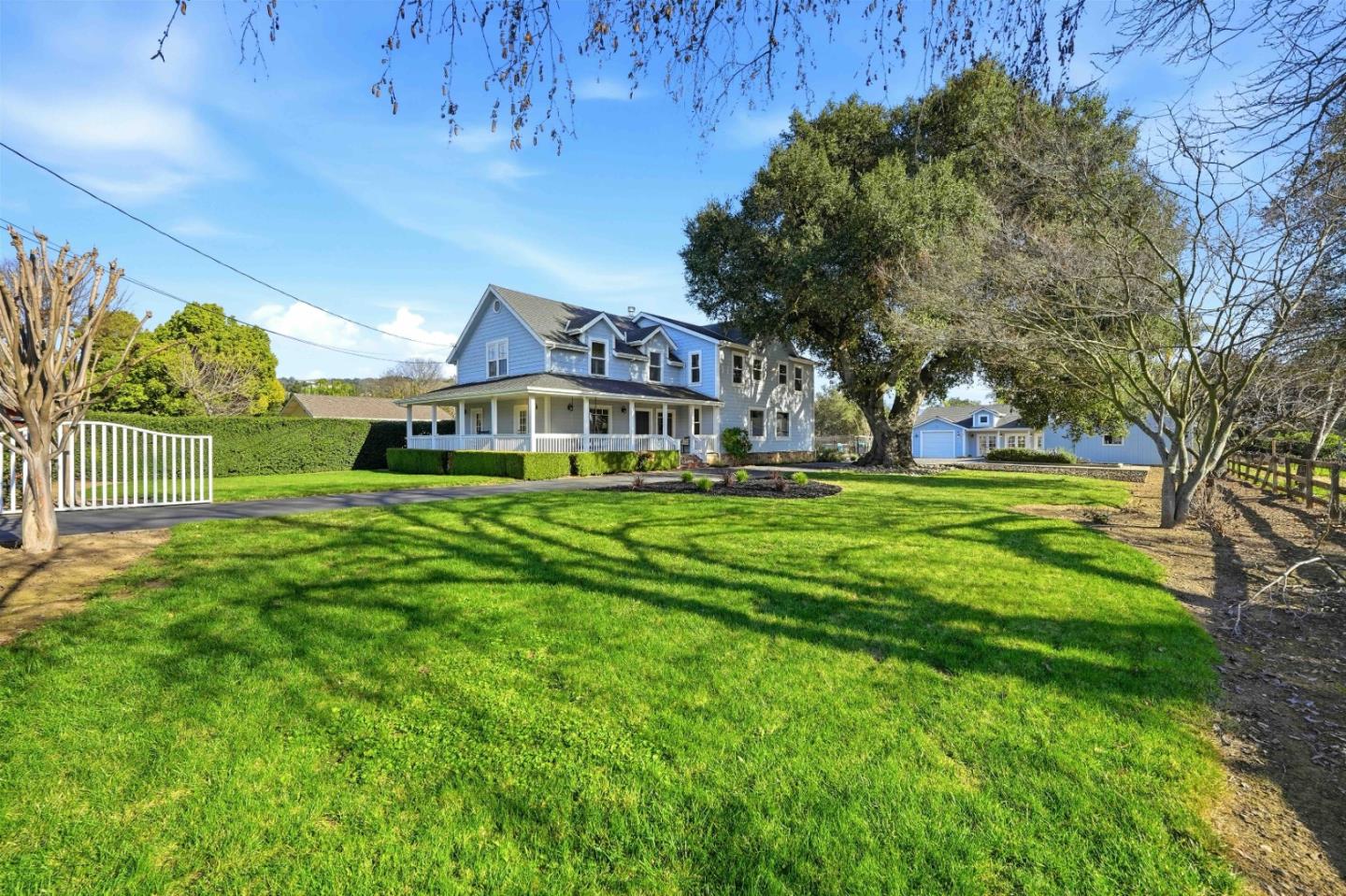 Blue two-story house with a wraparound porch, broad lawn, white driveway gate, and detached garage.