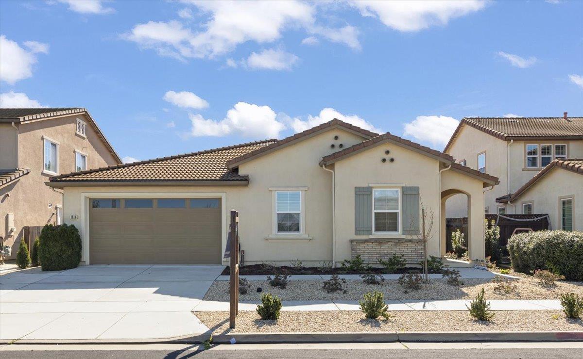 Single-story beige home with a tile roof, two-car garage, and gravel front yard.
