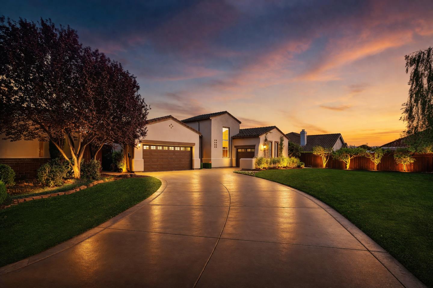 Stucco home with a curved driveway and illuminated landscaping beneath a colorful sunset sky.