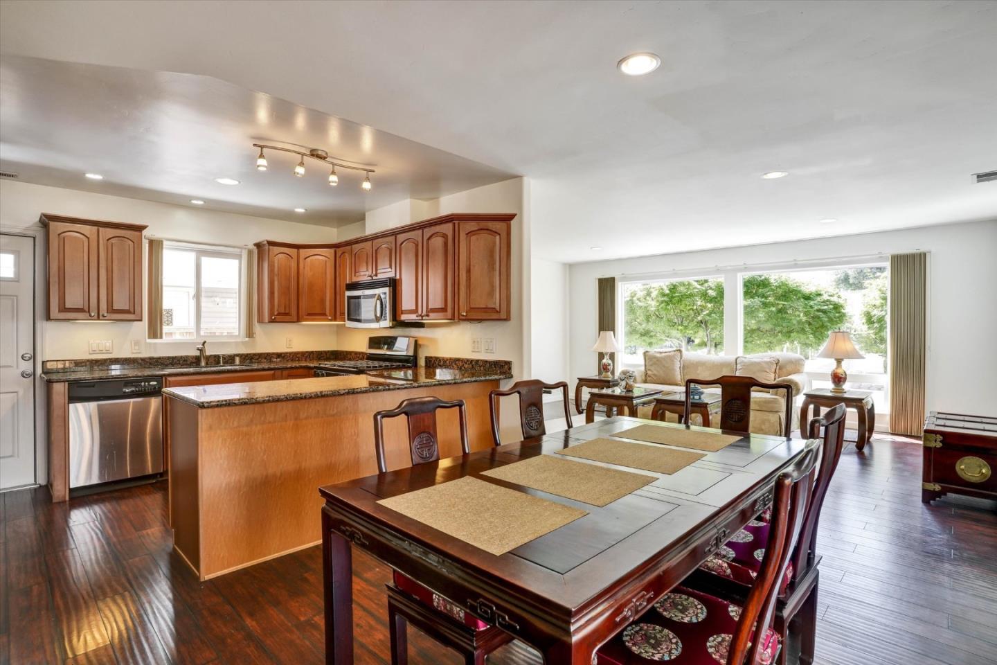 Open-plan kitchen and dining room with wood cabinets, granite counters, and a large dining table beside broad windows.