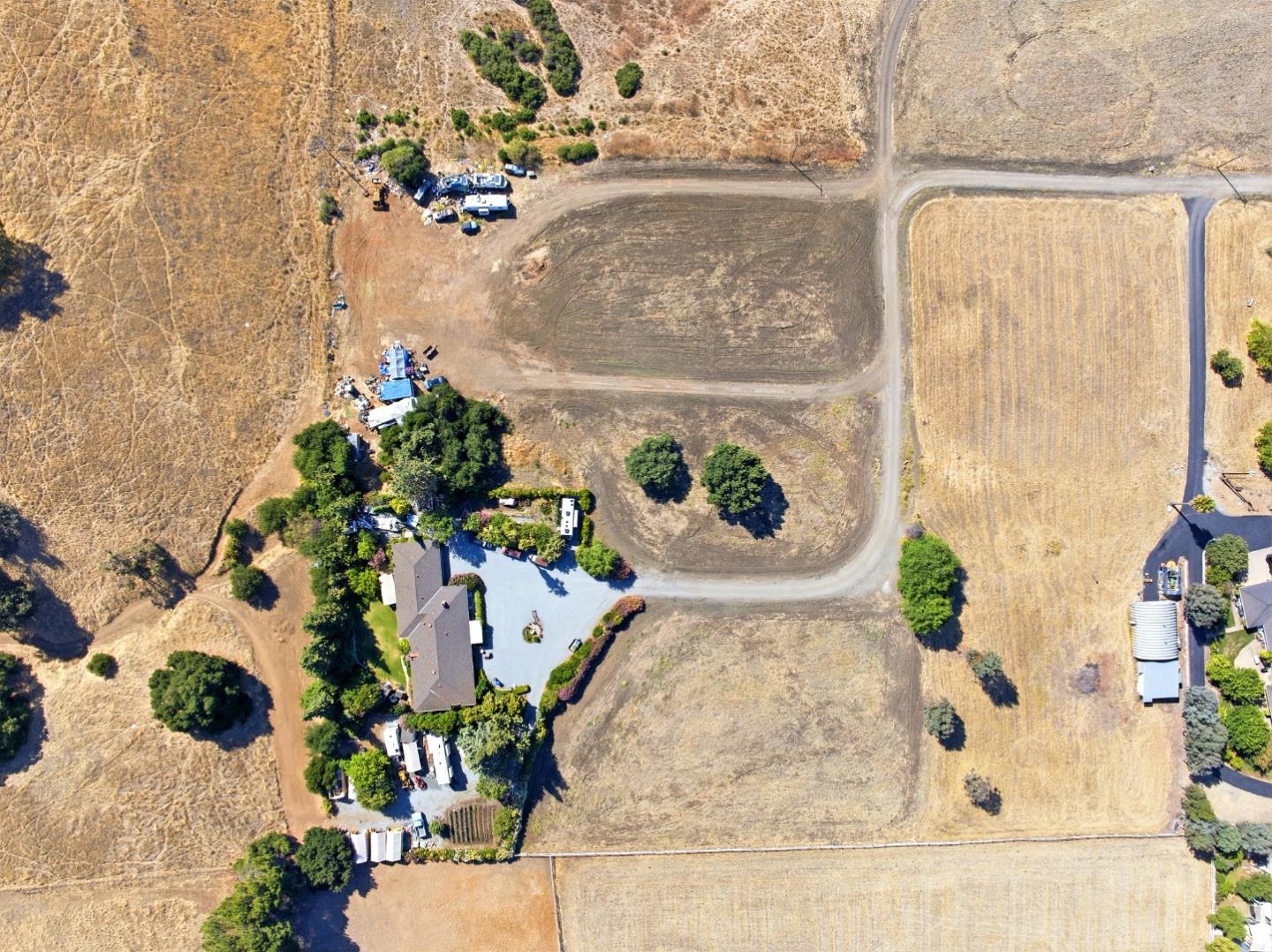 Aerial view of dry open land with dirt roads and buildings beside a cluster of trees.