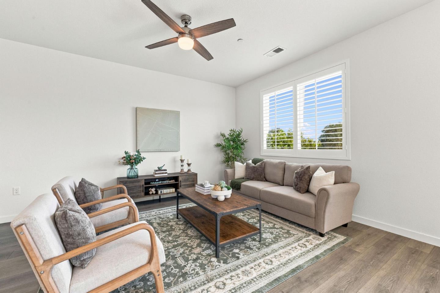 Staged living room with a beige sofa, wood-frame chairs, patterned rug, and large shuttered window.