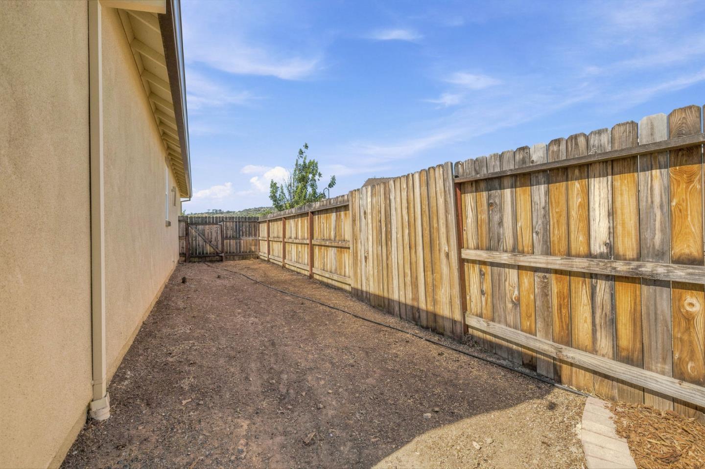 Fenced side yard beside a beige stucco home, with bare ground and a tall wooden privacy fence.