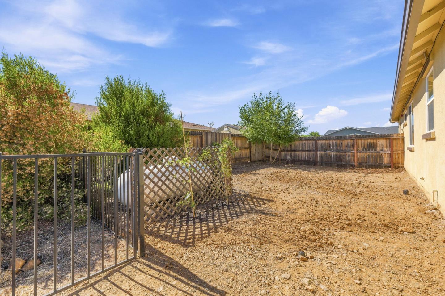 Fenced backyard with dry ground, a metal gate, lattice divider, trees, and wooden perimeter fence.