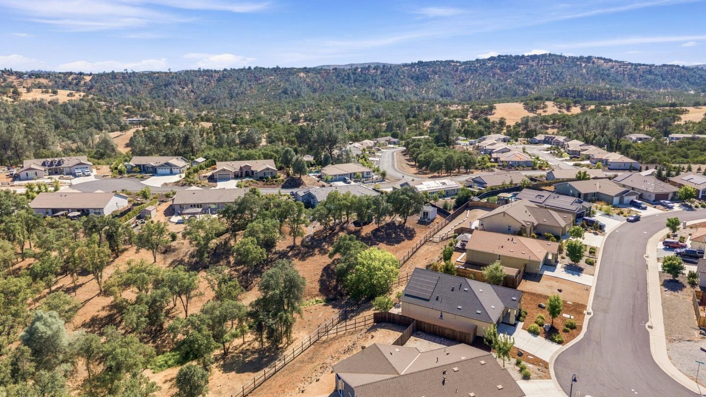 Aerial view of homes along a curving street beside wooded, dry terrain and rolling hills.