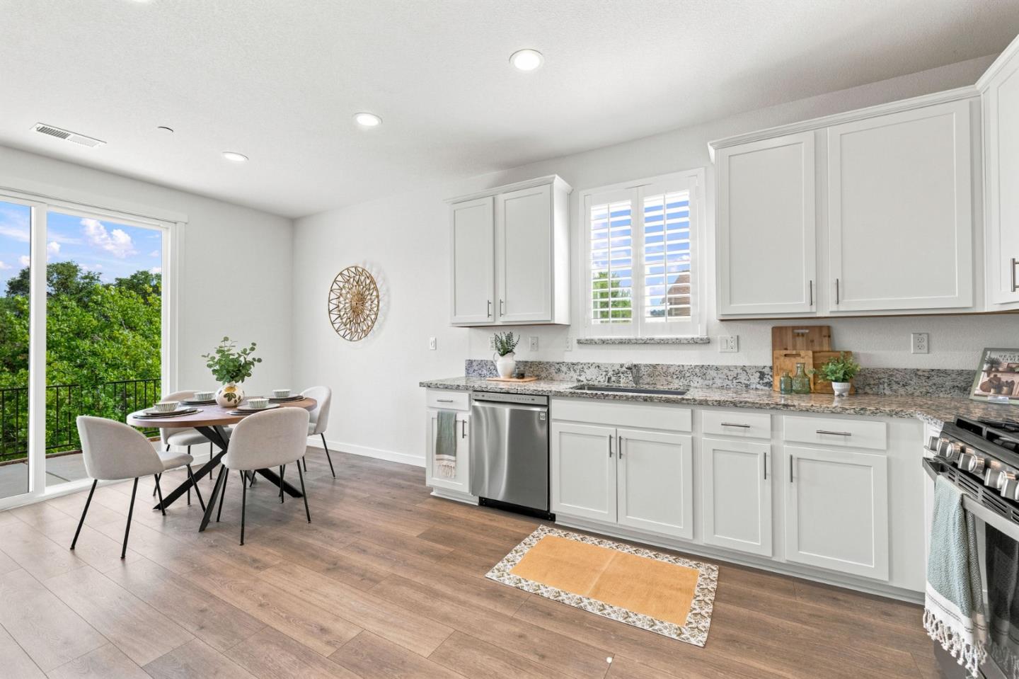 Bright white kitchen with granite counters, stainless steel appliances, and a dining nook beside leafy trees.