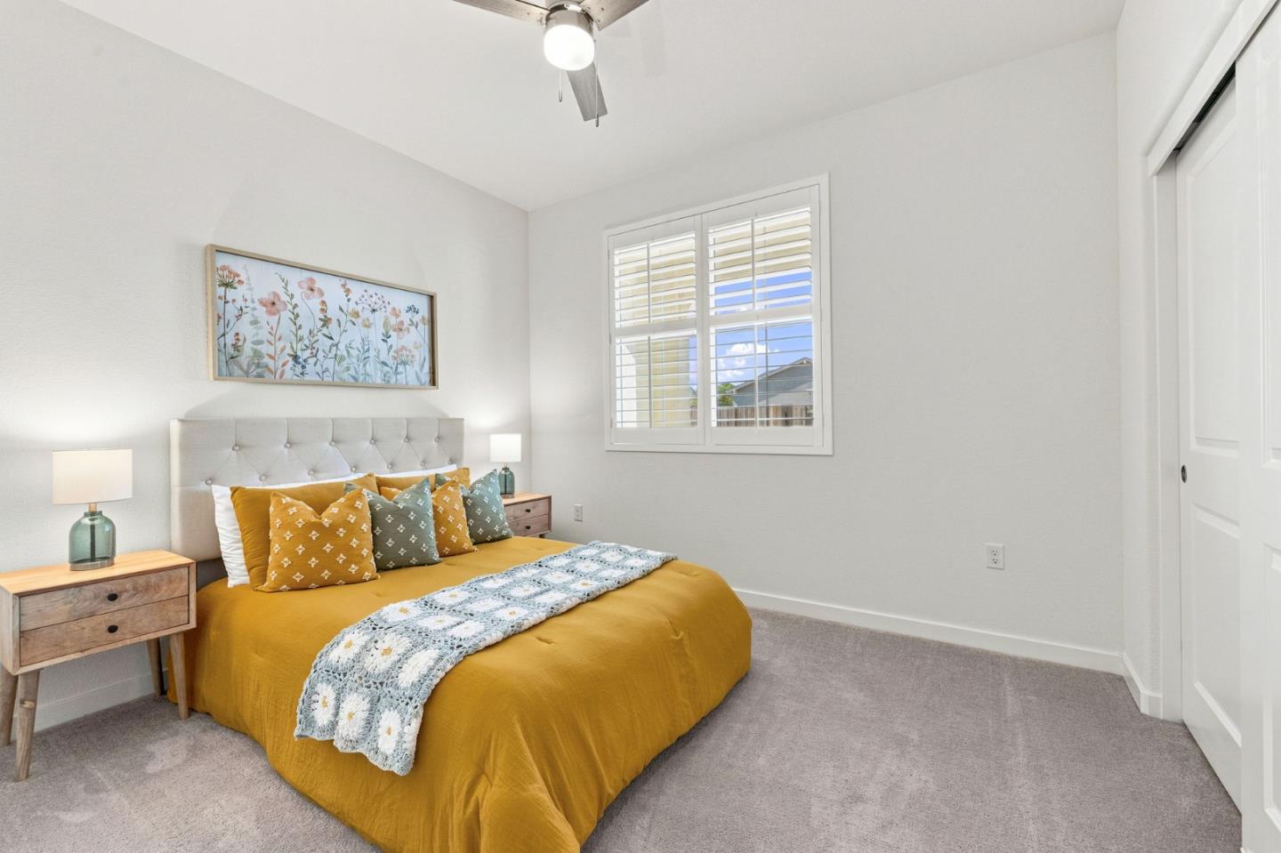 Staged bedroom with a mustard-yellow bedspread, patterned pillows, and a shuttered window.