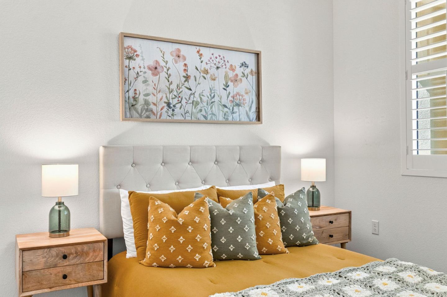 Bedroom with a mustard-yellow bedspread, patterned pillows, and floral wall art above a tufted headboard.