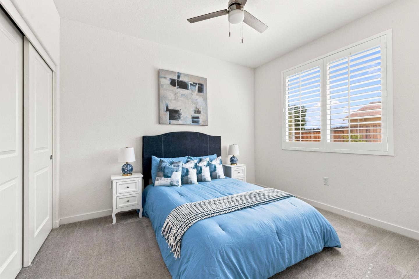 Bright bedroom with a blue bed, dark headboard, white nightstands, and plantation shutters.