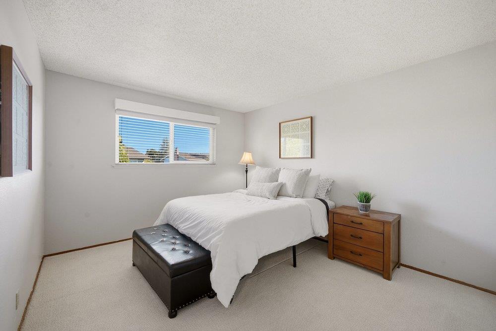 Bedroom with a white bed, dark upholstered bench, and wide window with blinds.