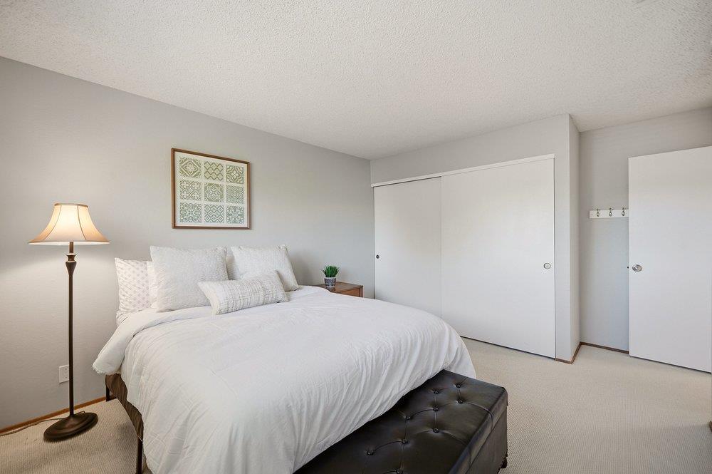 Staged bedroom with a white bed, dark tufted bench, floor lamp, and sliding closet doors.