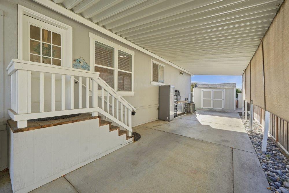Covered concrete patio beside a light-colored home, with white steps and a storage shed.