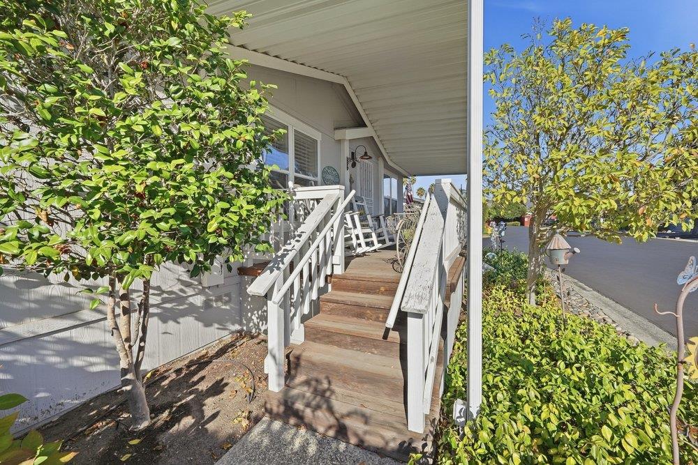 Home’s covered front porch with wooden steps, white railings, and rocking chairs beneath leafy trees.
