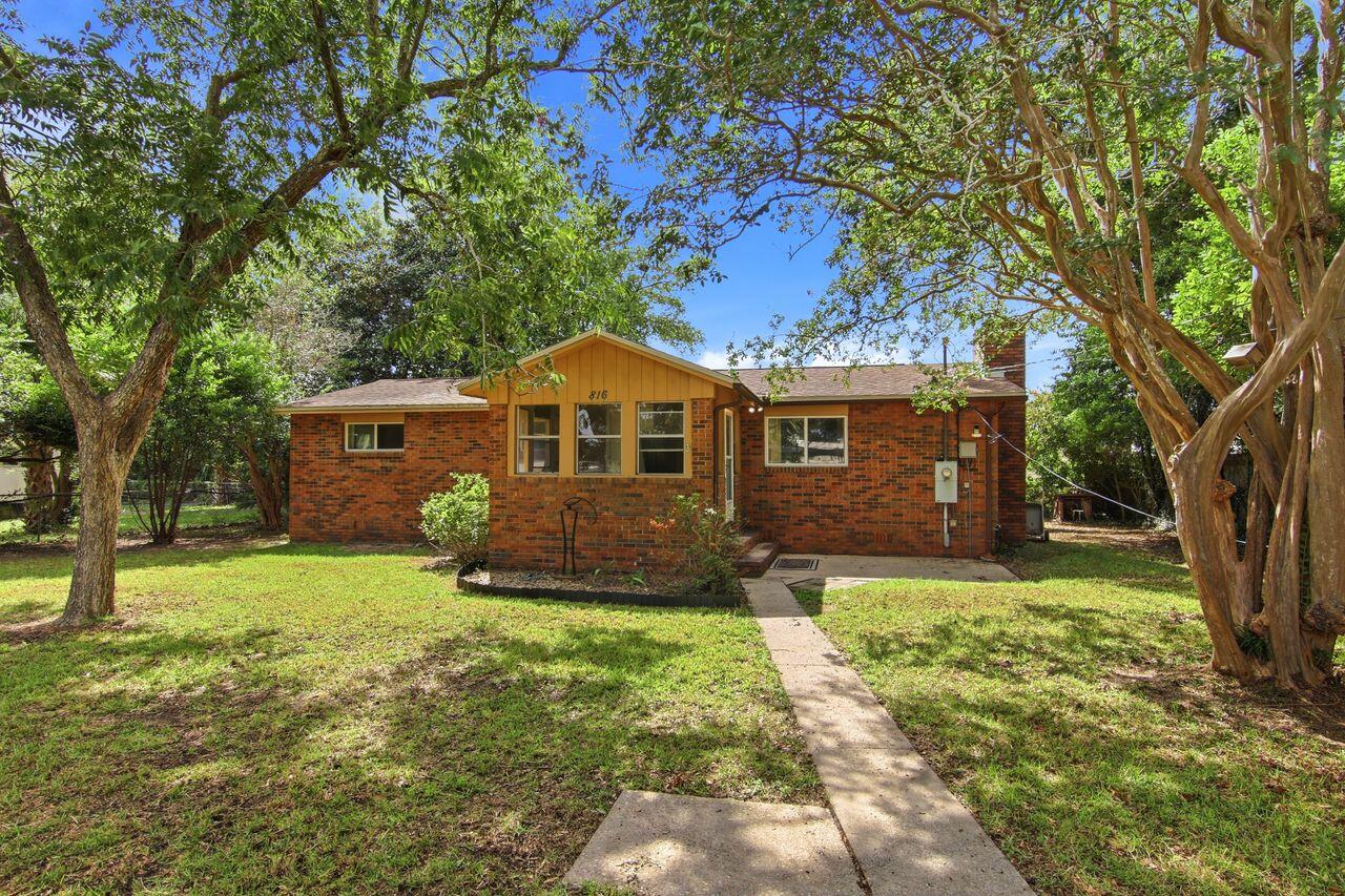 Single-story brick house with a tan front gable, shaded by mature trees and reached by a concrete walkway.