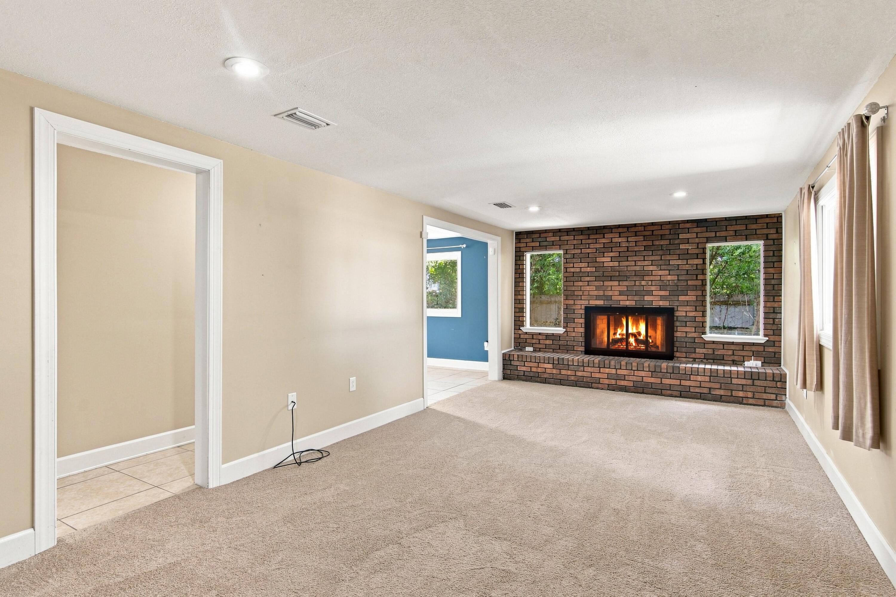 Empty living room with a brick fireplace, beige carpet, and windows along two walls.