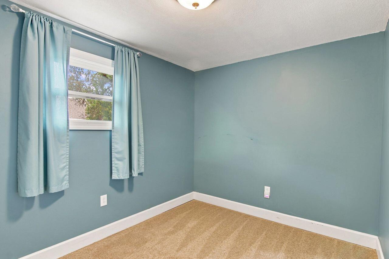 Empty bedroom with blue-green walls, tan carpet, and light blue curtains around the window.