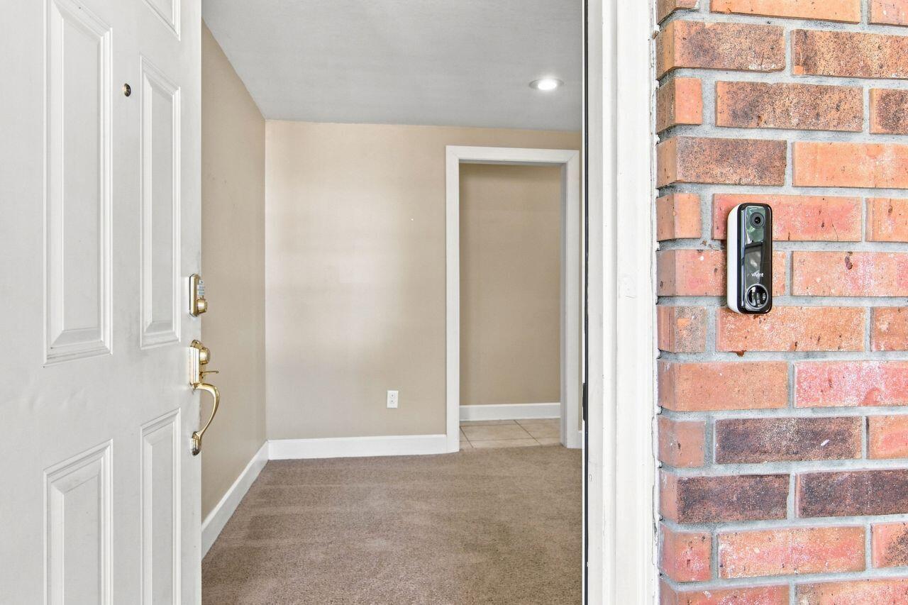 Vacant entry foyer with beige walls, carpeted floor, and a video doorbell beside the open front door.