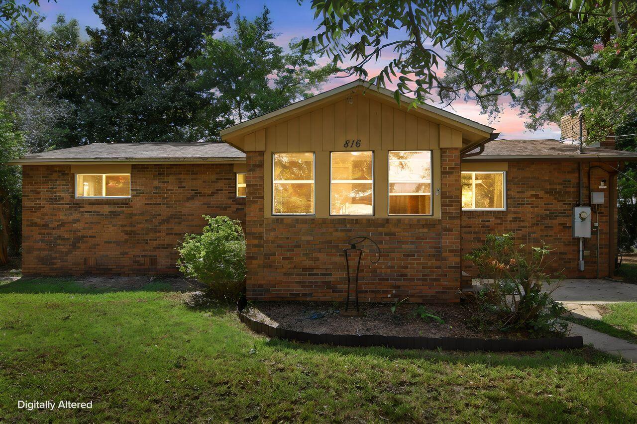 Brick single-story home with a projecting gabled entry, lit windows, and mature trees.