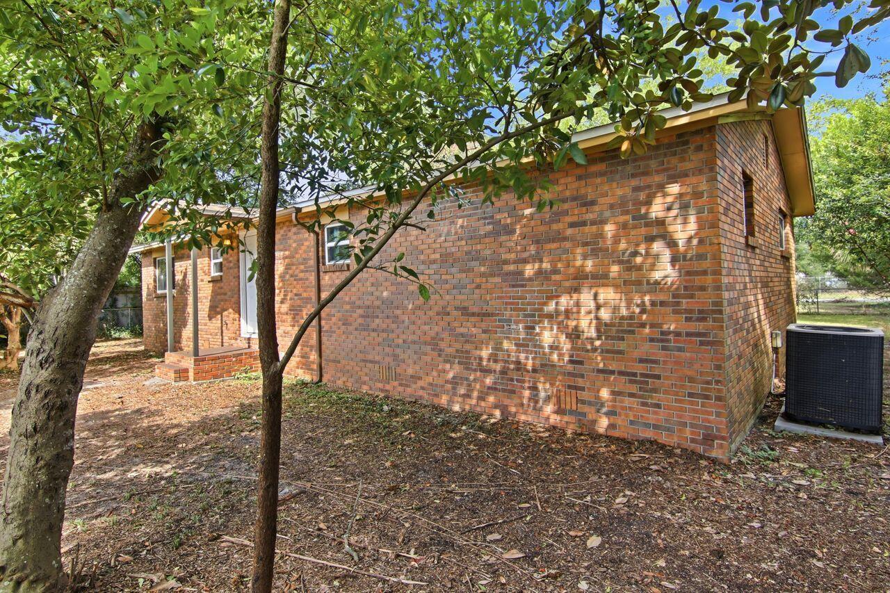 Side of a single-story brick house shaded by trees, with a white door and outdoor air-conditioning unit.