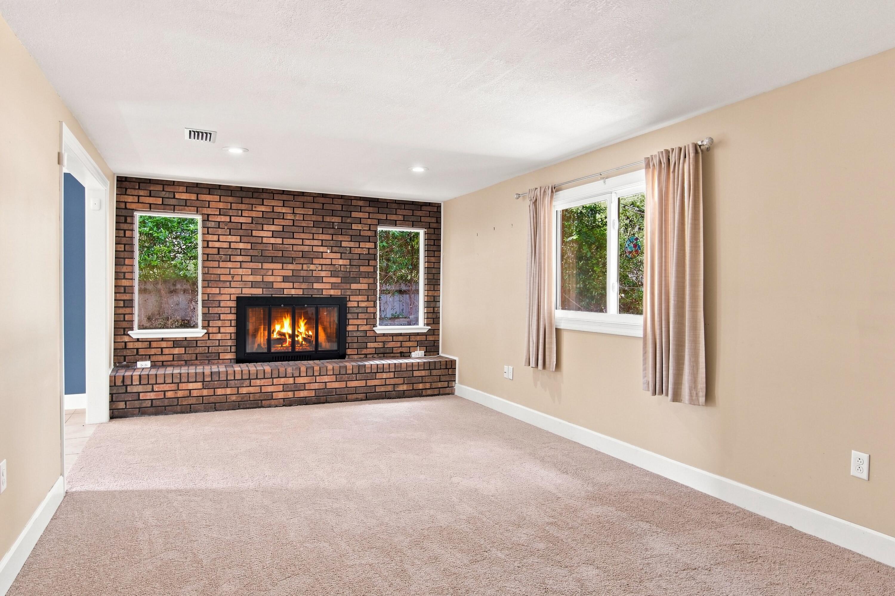 Empty living room with a lit brick fireplace and beige carpet.