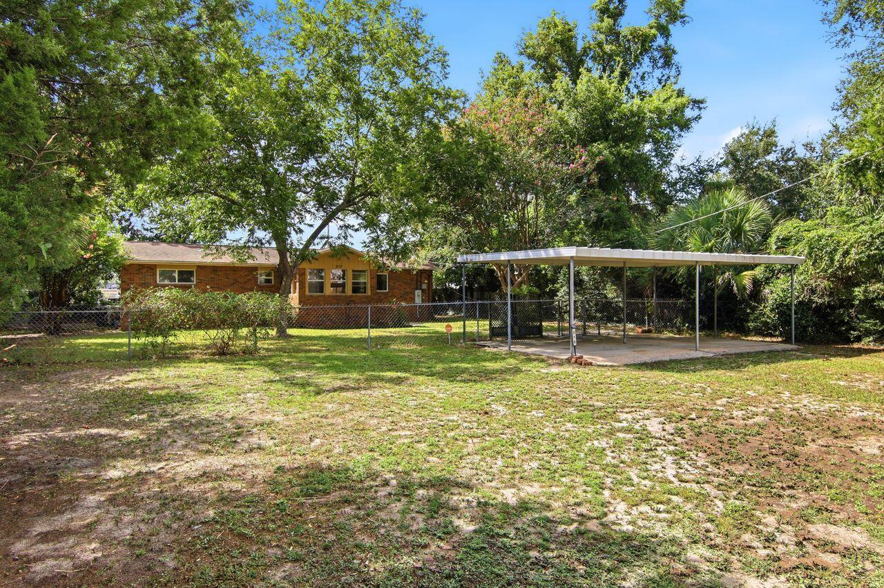 Single-story brick house behind a chain-link fence, with a broad carport and mature trees.