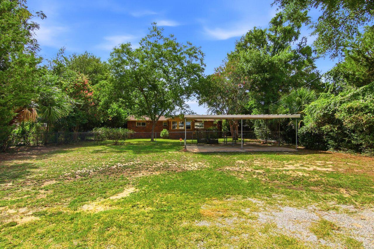 Single-story brick house behind a fenced, tree-lined yard, with a long covered carport.