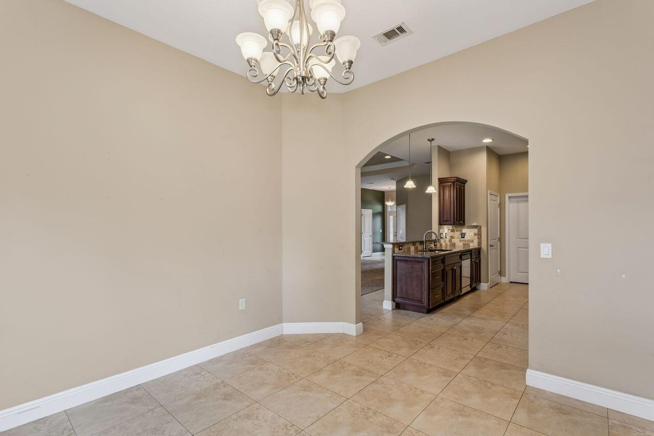 Empty tiled dining area with a chandelier and arched opening to a kitchen with dark wood cabinets.