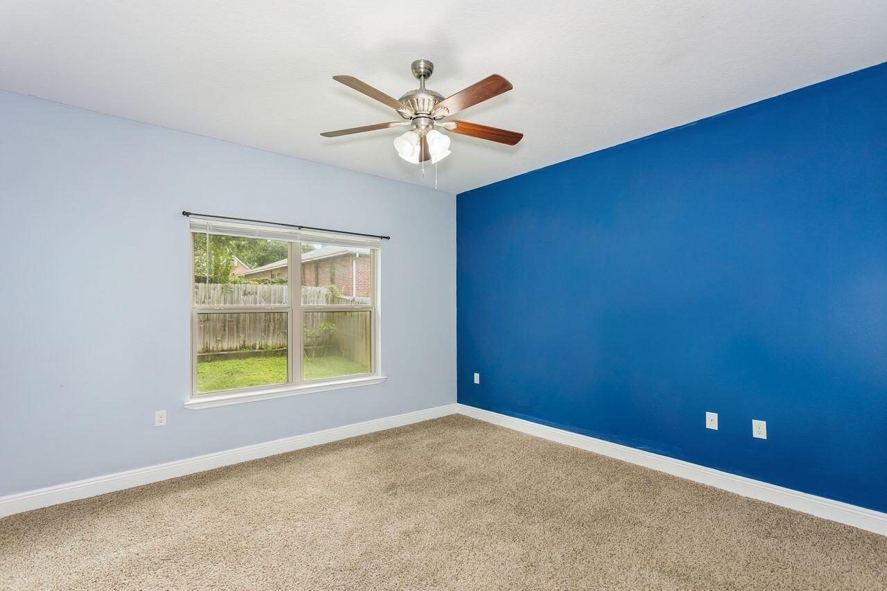 Empty bedroom with a blue accent wall, beige carpet, ceiling fan, and window overlooking a fenced yard.