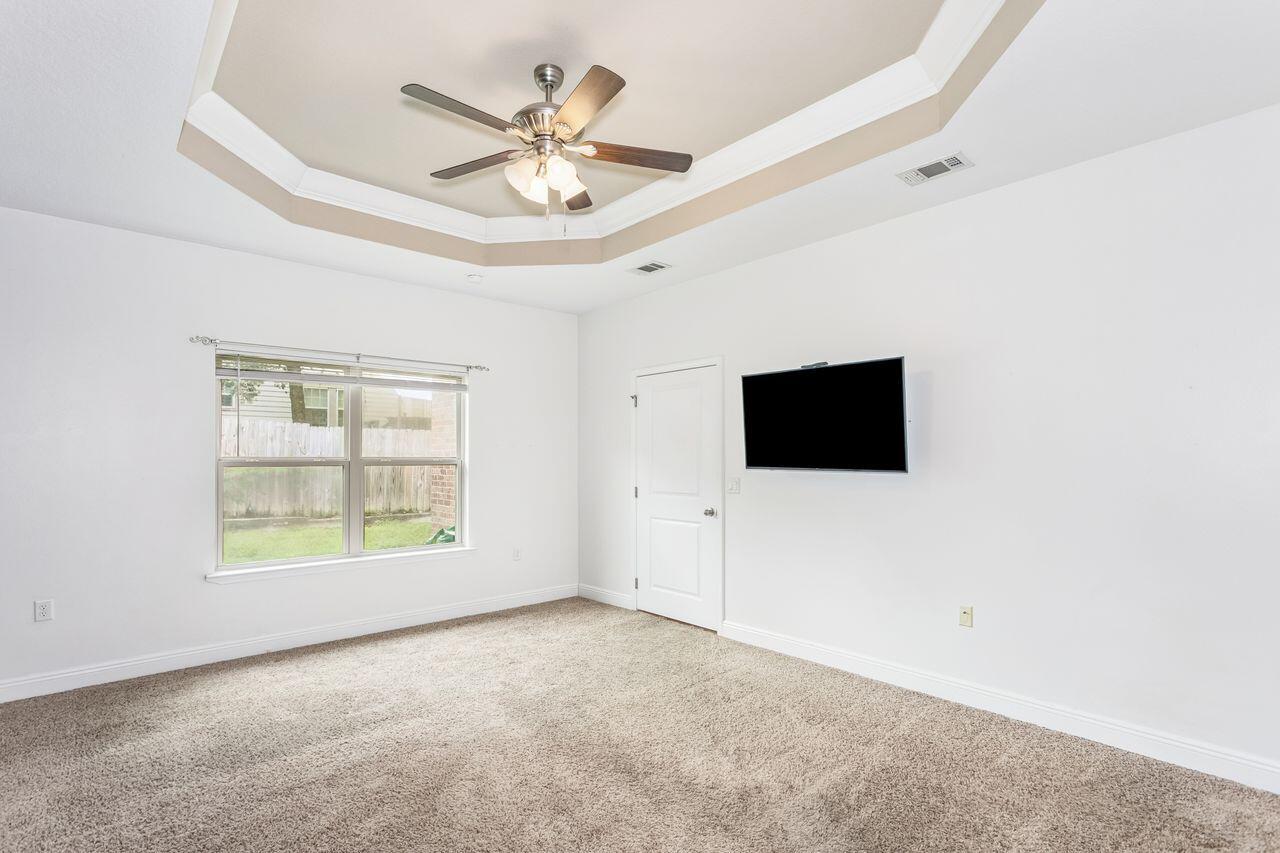 Empty carpeted bedroom with a tray ceiling, ceiling fan, window, and wall-mounted television.