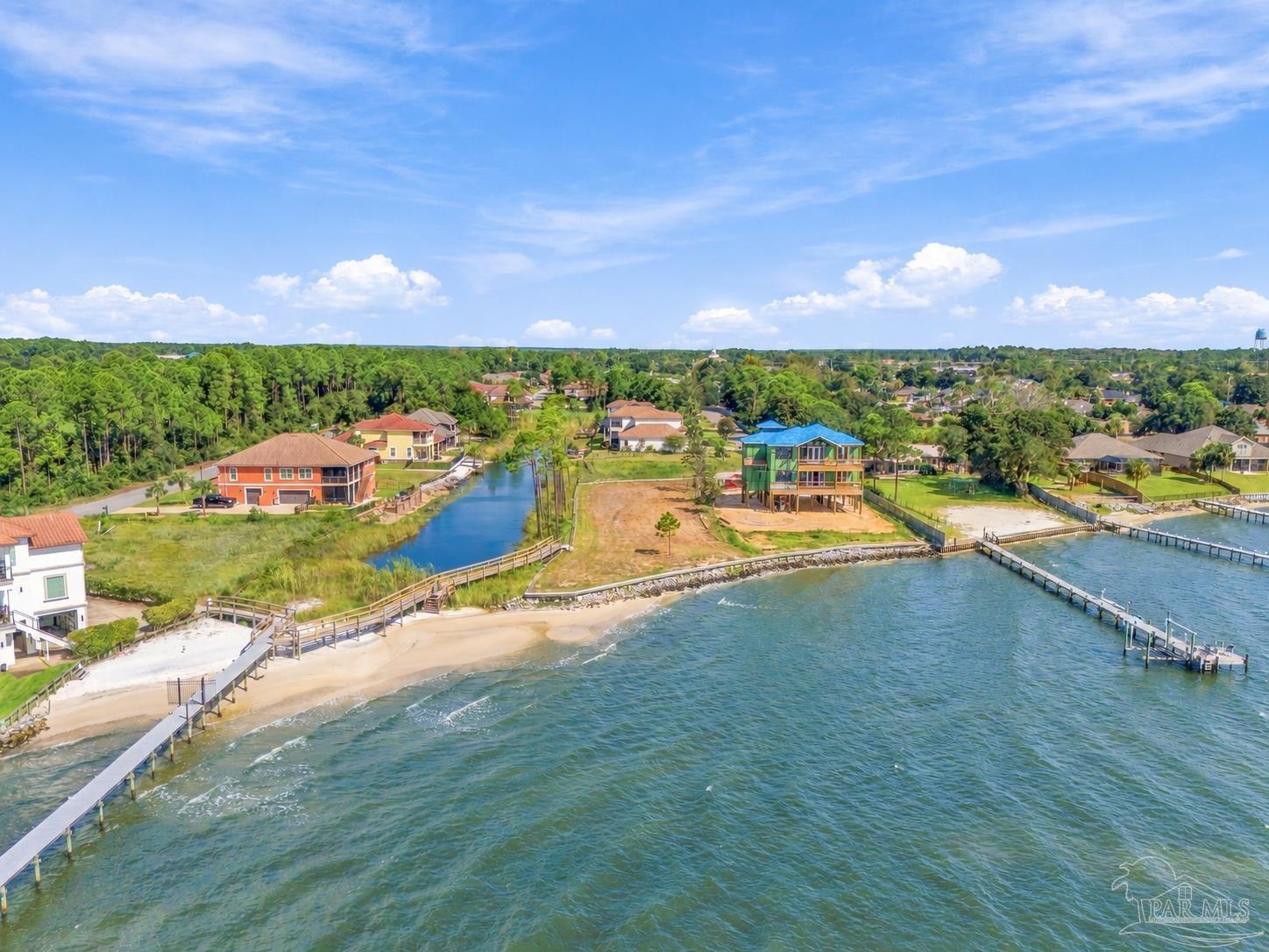 Waterfront home under construction on a sandy lot, with docks and neighboring houses along the shore.