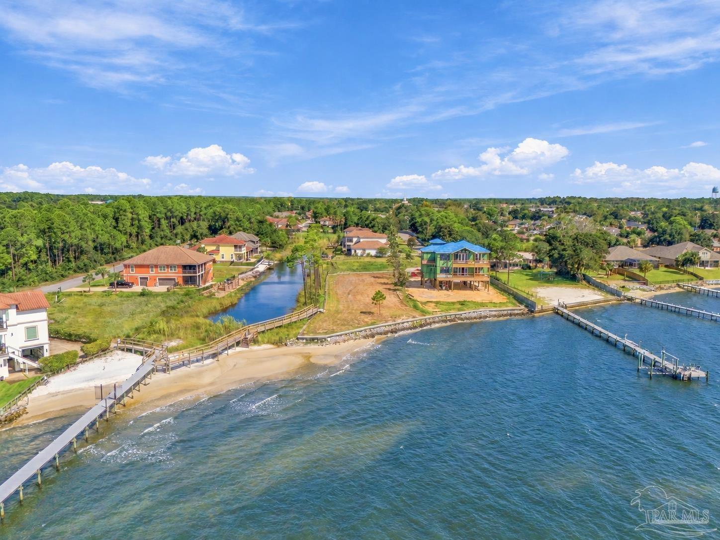 Waterfront home under construction beside a sandy shoreline, docks, and neighboring houses.