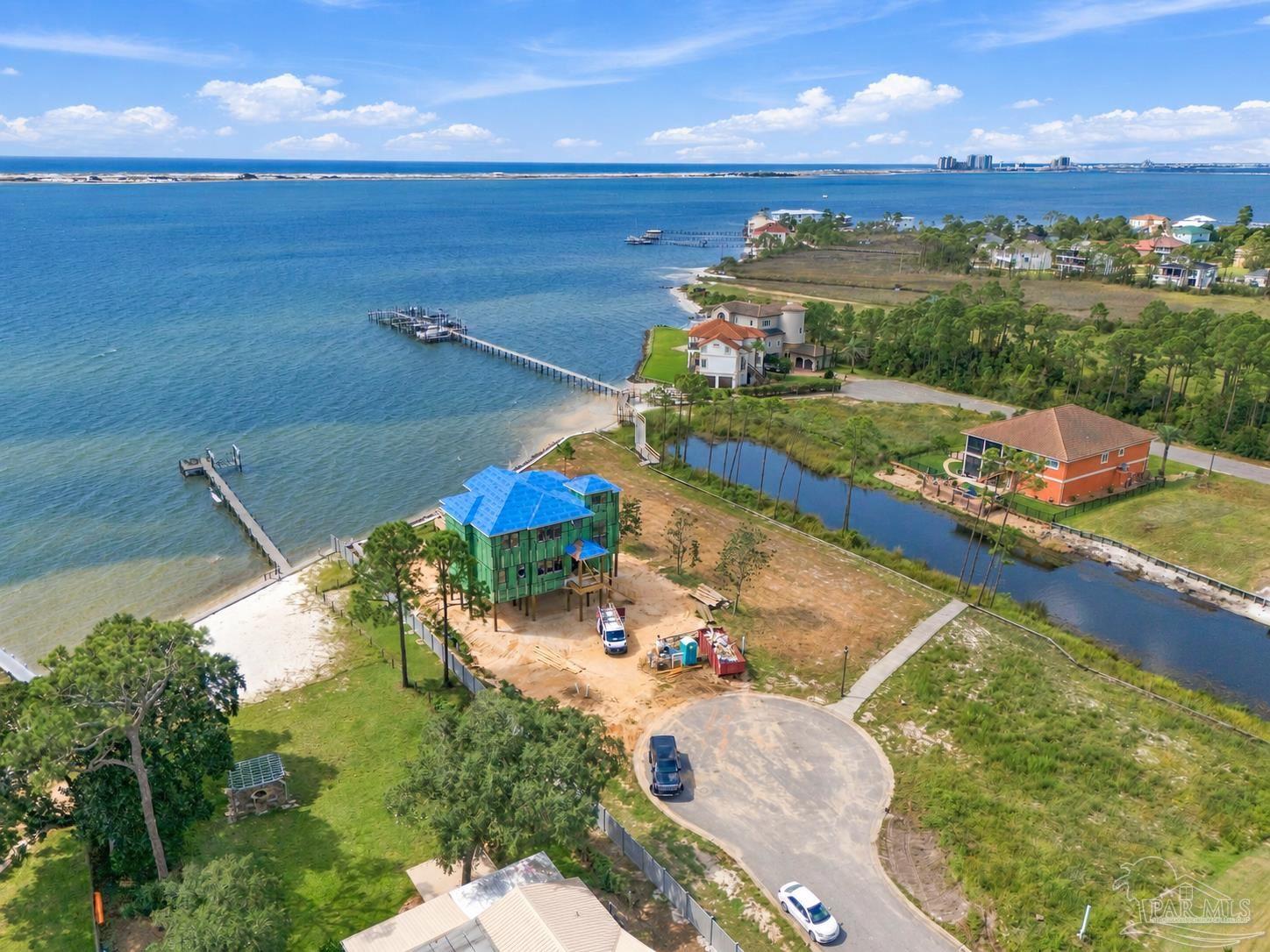 Aerial waterfront view of a house under construction, with a sandy shoreline and docks extending into the bay.