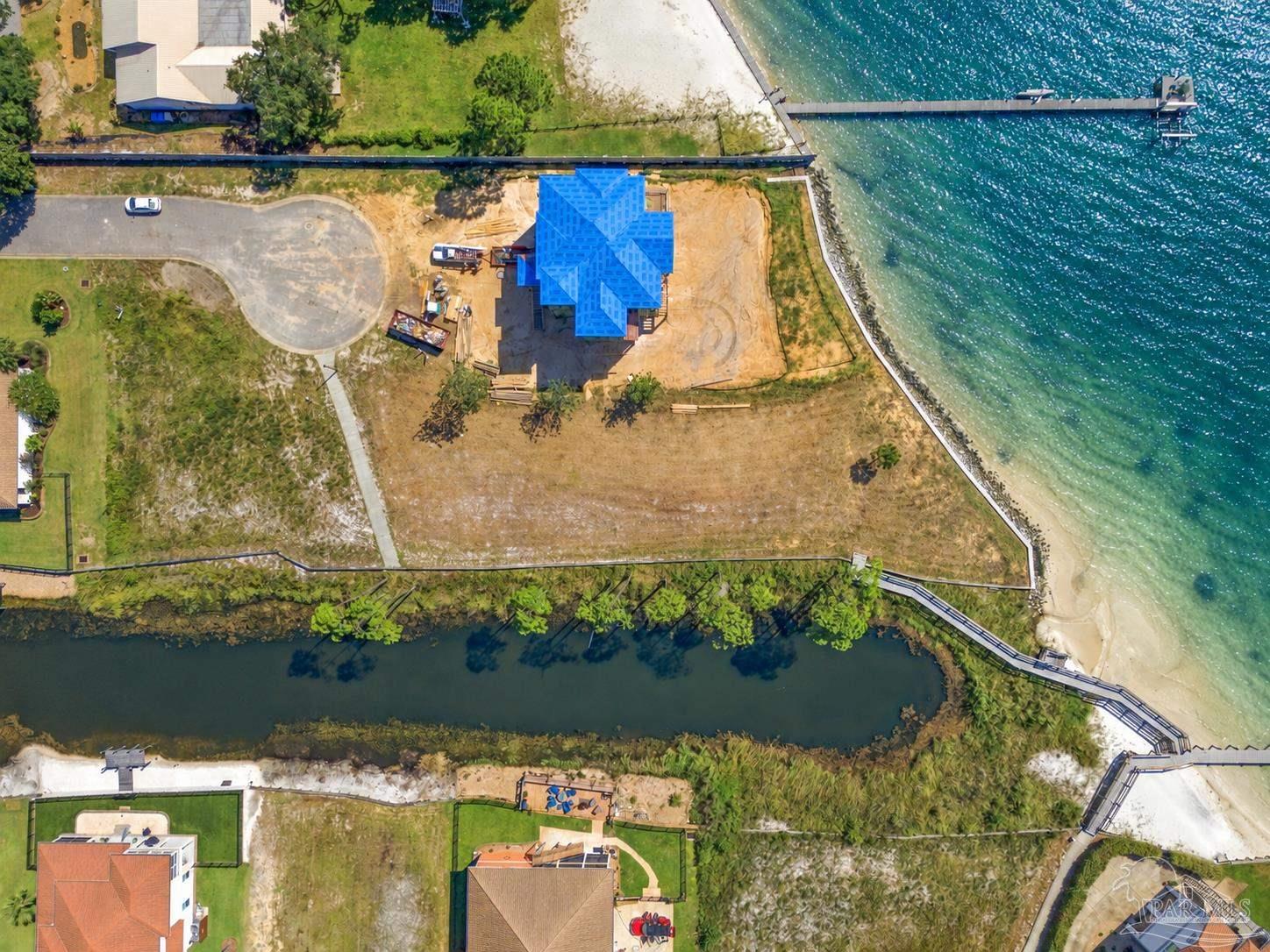Aerial view of a waterfront house under construction, with a dock and turquoise water beside the lot.