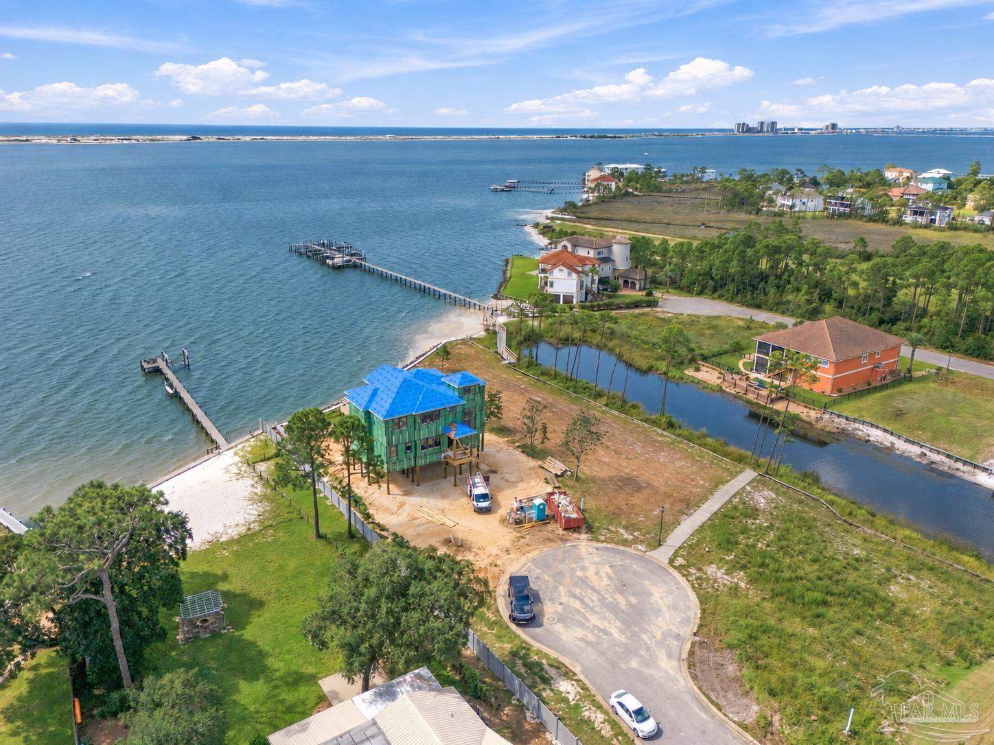 Waterfront home under construction with a blue roof, shoreline, and docks beside a broad bay.
