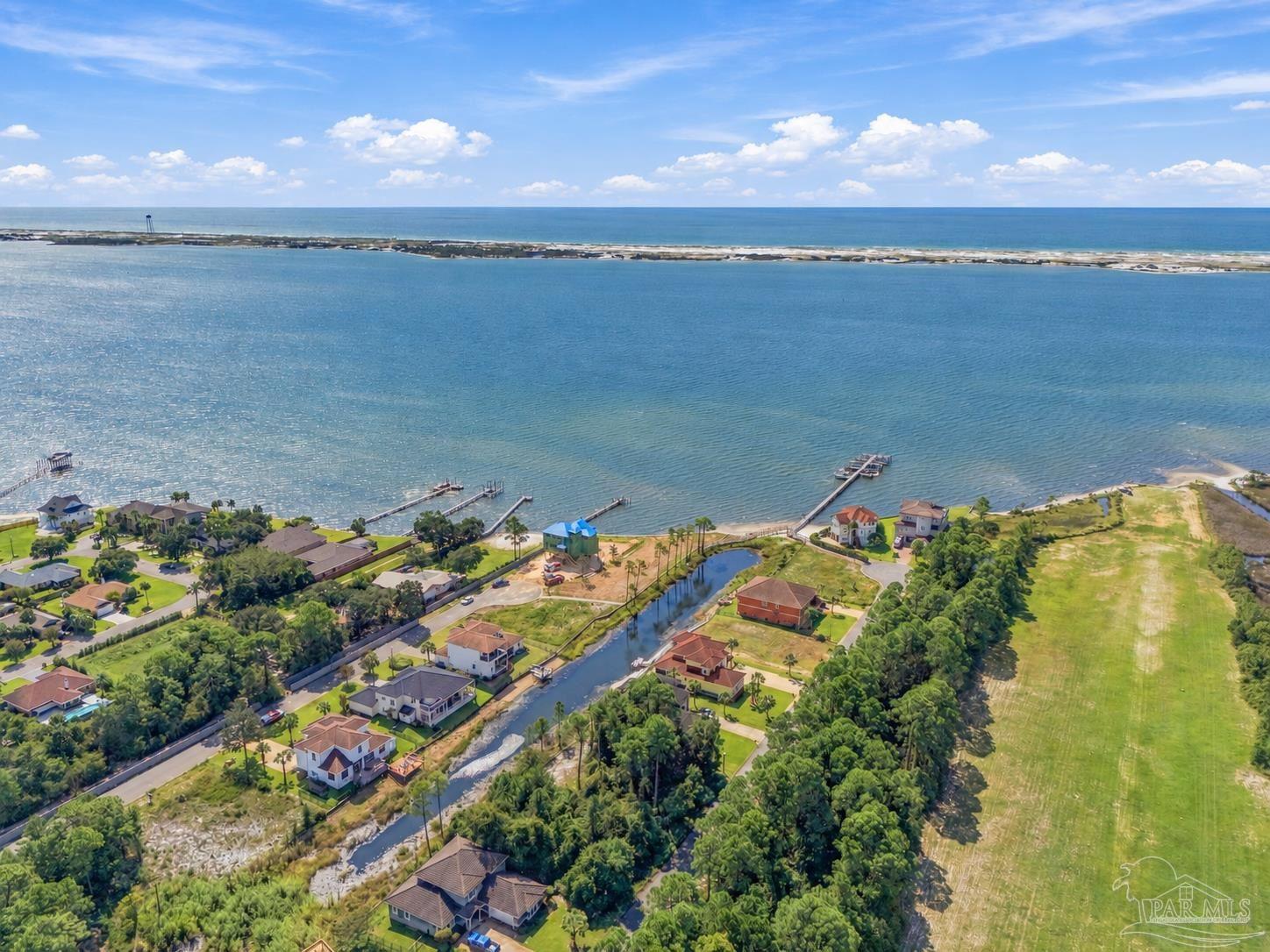 Aerial view of waterfront homes and canal beside a broad bay, with docks and a sandy barrier shoreline.