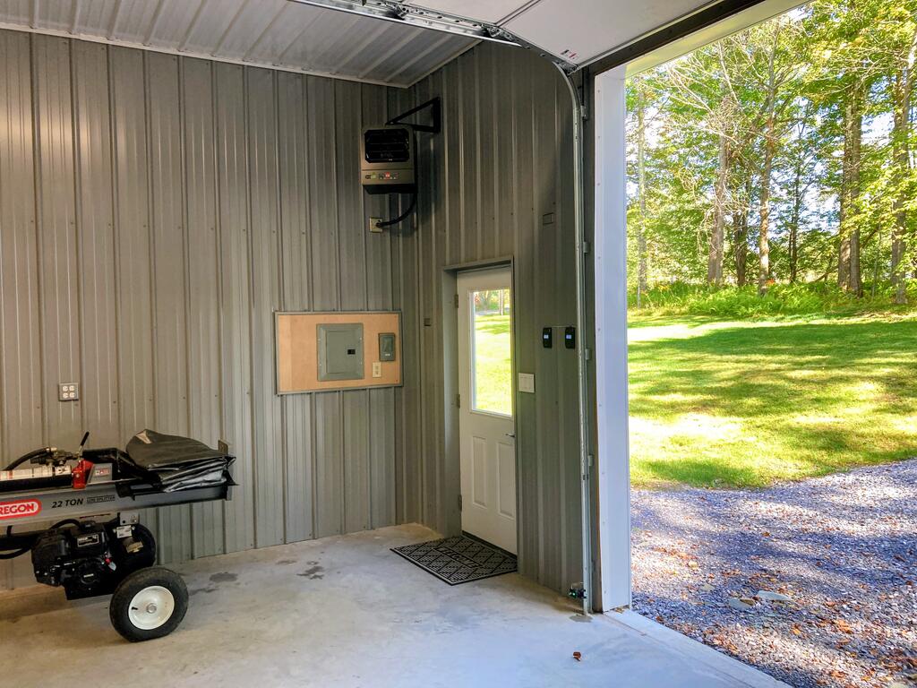 Metal-sided garage with an open overhead door facing a sunny lawn and trees.