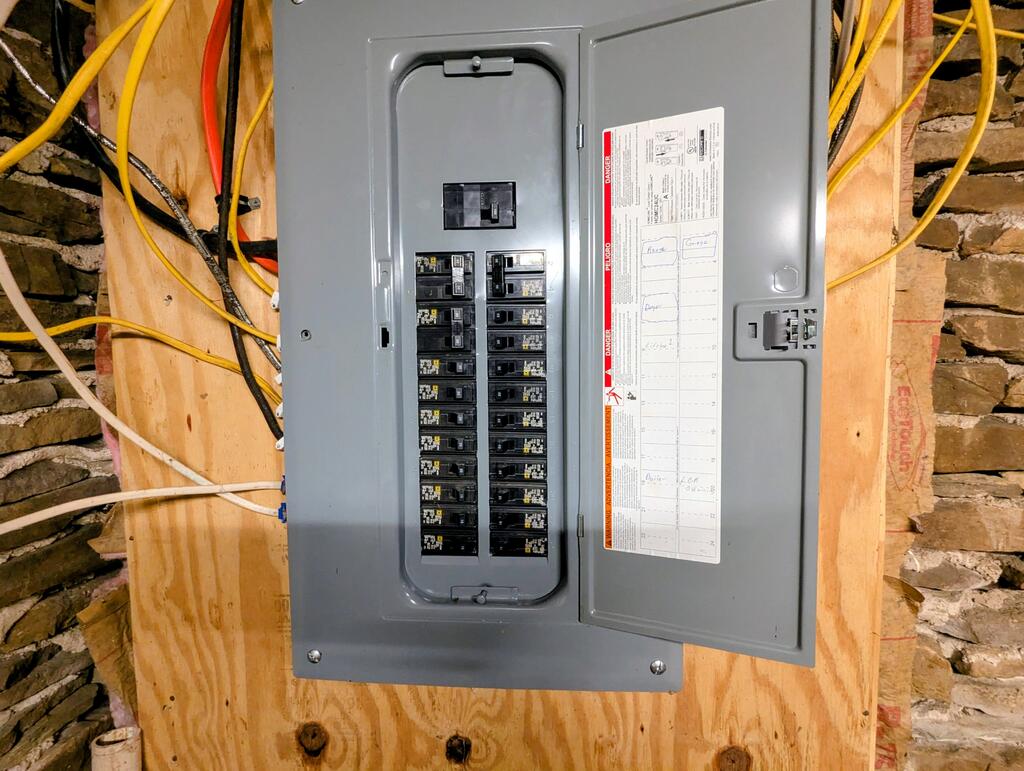 Open breaker panel mounted on plywood, with exposed wiring and stone walls.