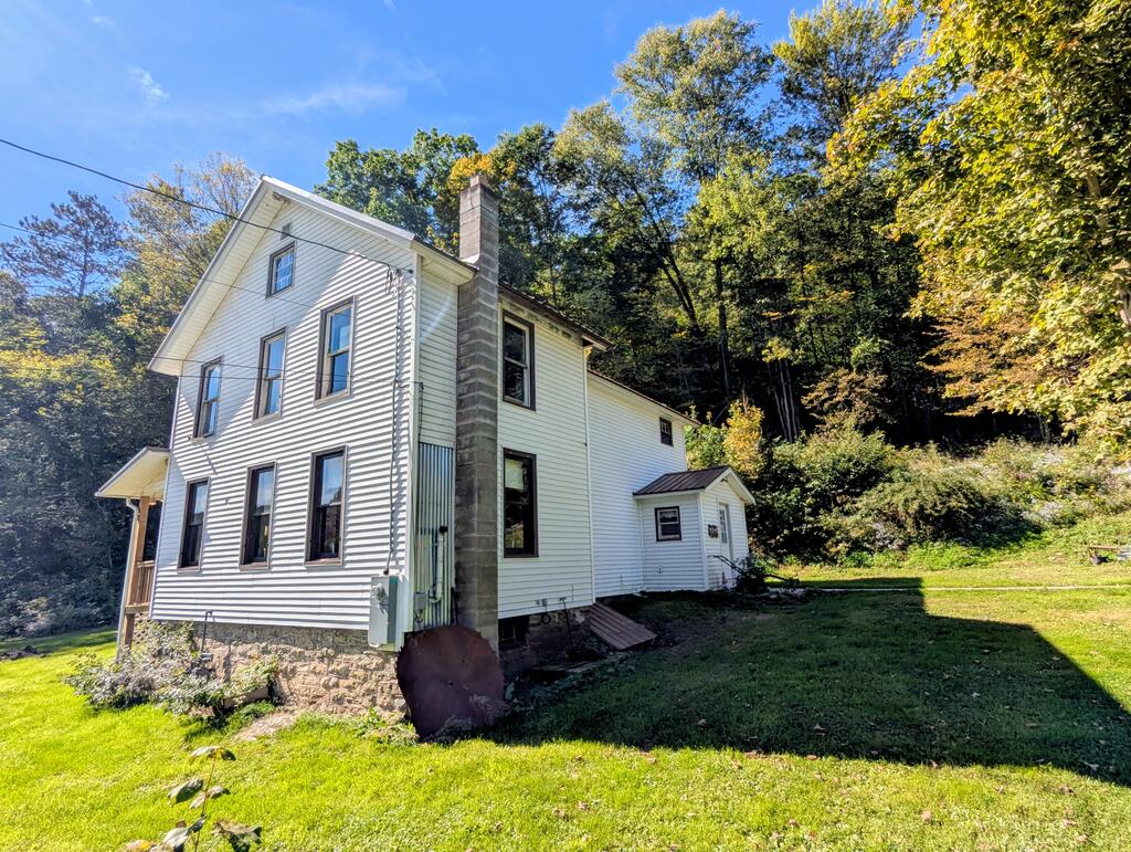 Two-story white-sided house with a tall stone chimney, lawn, and wooded hillside.