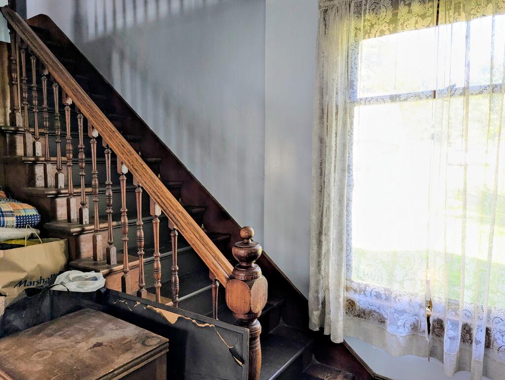 Wooden staircase with turned balusters and carved newel post beside a bright lace-curtained window.