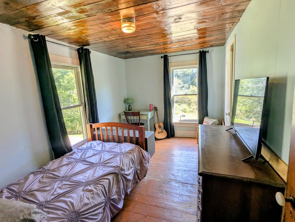 Bedroom with a wood-plank ceiling, hardwood floor, and windows overlooking trees.