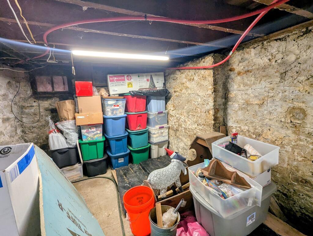 Stone-walled basement storage area with stacked bins, boxes, and exposed ceiling pipes.