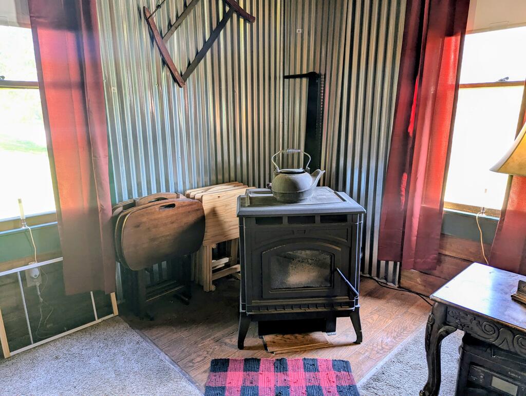 Rustic room with a wood-burning stove, kettle, corrugated metal walls, and folding chairs.