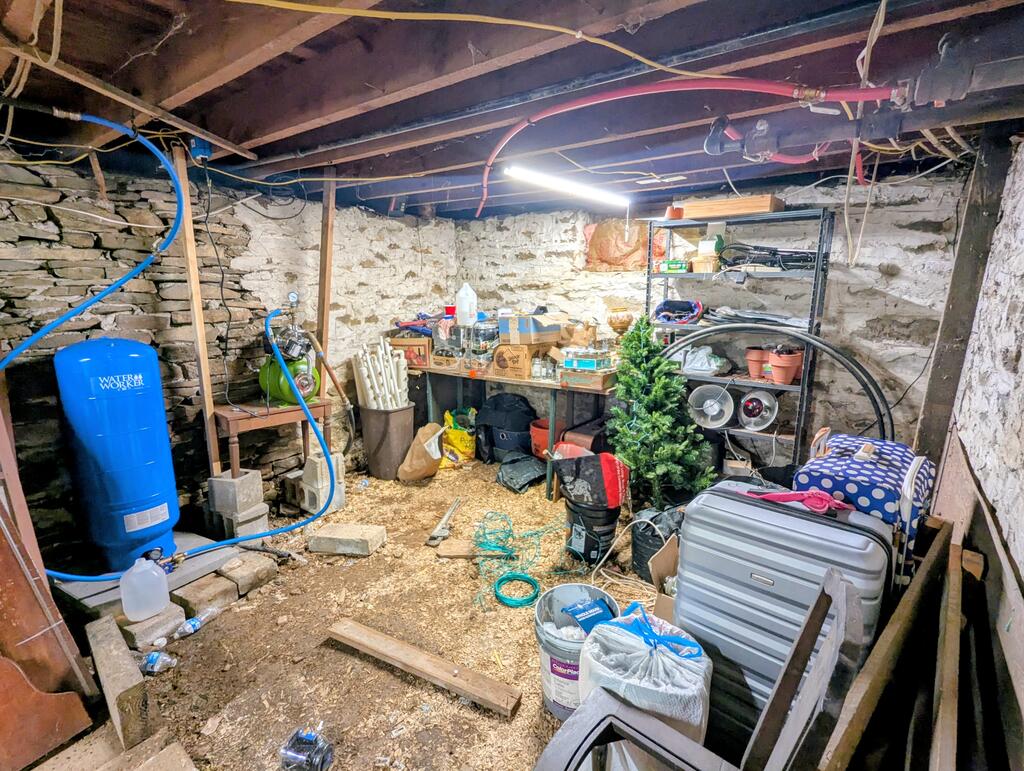 Basement with rough stone walls, exposed joists, a blue water softener, and crowded storage shelves.