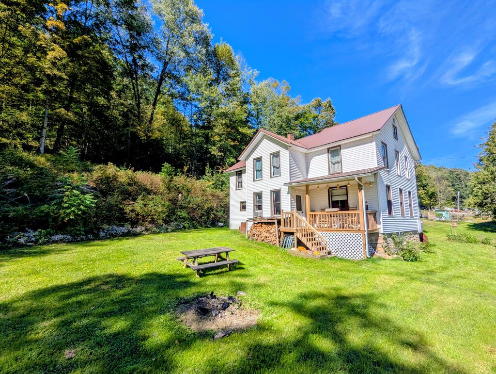 White two-story house with a red roof and covered porch beside a wooded hillside.