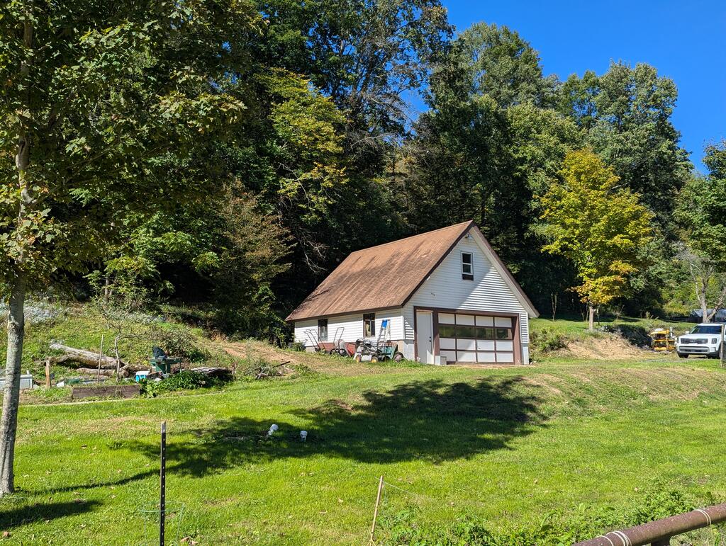 White detached garage with a brown gabled roof in a grassy, wooded clearing.