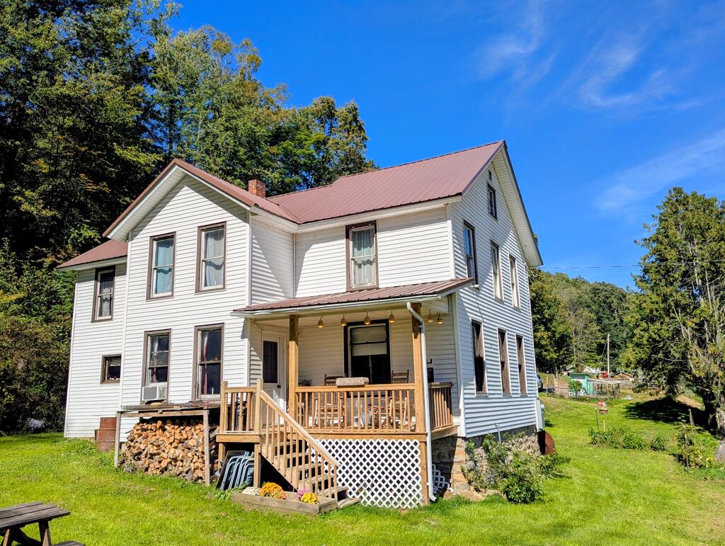 White two-story house with a red metal roof, covered wooden porch, and wooded lawn.